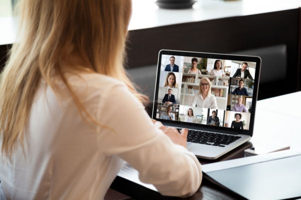 View over businesswoman shoulder sit at workplace desk looks at pc screen during group videocall with different age and ethnicity colleagues using application and webcam, distant communication concept